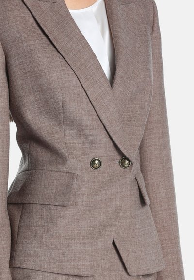 Gros plan sur un blazer croisé taupe avec des revers pointus, deux boutons décoratifs, des poches à rabat, et un top blanc en dessous.