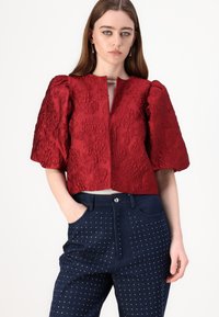 Red textured cropped jacket with puff sleeves and a round neckline. Paired with navy blue pants featuring white polka dots and pockets.