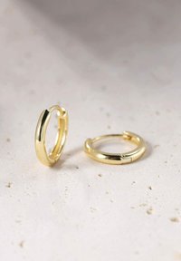 Orecchini a cerchio in oro con una finitura liscia e lucida. Presentano un design semplice e un meccanismo di chiusura sicuro. Due orecchini sono mostrati su uno sfondo chiaro.