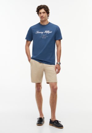 Mand stående iført en blå Tommy Hilfiger t-shirt, beige shorts, sorte sko og et ur på håndleddet mod en ensfarvet hvid baggrund.