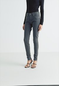 Jeans skinny gris con un corte entallado, diseño de cinco bolsillos y cierre frontal con botón, combinados con unos tacones altos negros con tiras.