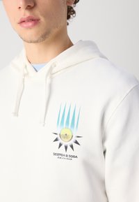 Valge kapuutsiga särk pehmest kangast, millel on kollase, musta ja sinise päikesegraafika ning allpool tekst "SCOTCH & SODA Amsterdam".