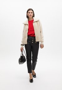 Beige Kunstfelljacke über einem roten Sweatshirt, kombiniert mit schwarzen Lederhosen mit goldenen Knöpfen. Eine schwarze Handtasche und Schuhe runden das Outfit ab.