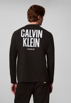 Homme vu de dos portant une chemise noire à manches longues avec un grand texte blanc "Calvin Klein Established 1968" dans le dos.