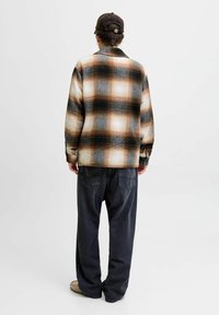 Plaid flannel shirt in brown, black, and cream tones, featuring a relaxed fit and a collar, paired with loose dark jeans and beige shoes.