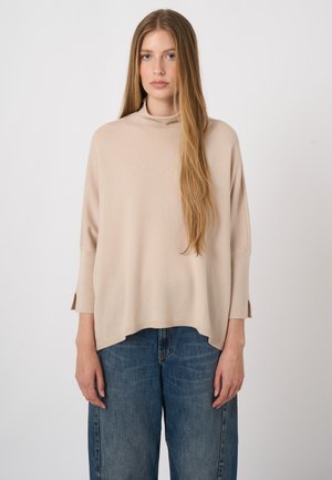Young woman with long straight hair wearing a loose beige turtleneck sweater and blue jeans standing against a plain light background.