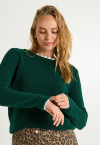 Pull en tricot vert avec un col rond agrémenté de dentelle blanche, manches longues et tissu texturé, associé à un pantalon imprimé léopard.