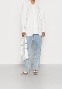 Camicia bianca a maniche lunghe con bottoni, jeans larghi di un blu chiaro, borsa a spalla bianca con tracolla borchiata e infradito bianchi.