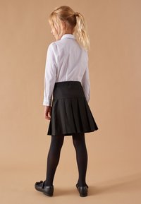 Schwarzer, plissierter Rock mit einem enganliegenden Taillenbund, kombiniert mit einem weißen Langarmshirt. Schwarze Strumpfhosen und Schuhe runden das Outfit ab.