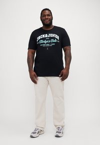 Čierne tričko s tyrkysovou grafikou "Jack & Jones Surfer's Club", okrúhly výstrih, nosené s svetlo béžovými nohavicami a béžovo-sivými teniskami.