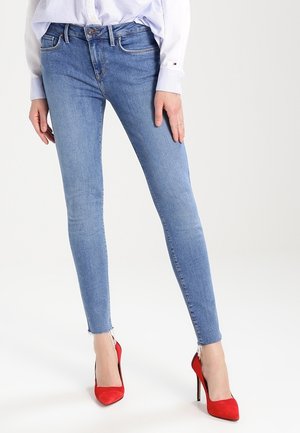 Personne portant un jean skinny bleu clair avec des ourlets effilochés, une chemise blanche boutonnée et des chaussures à talons rouges, debout devant un fond uni.