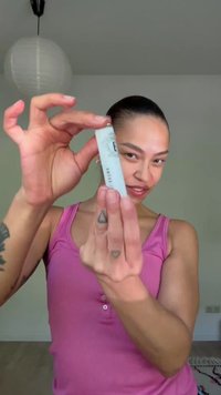Personne en débardeur rose tenant et montrant un petit tube de produit de soin de la peau ou cosmétique avec des tatouages visibles sur les doigts et le bras.