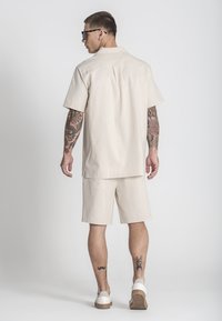 Camisa de manga corta beige y pantalones cortos a juego, ambos en una tela suave. Incluye un pliegue en la parte posterior, bolsillos laterales y zapatillas blancas.