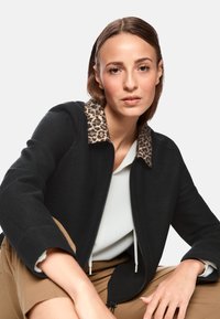 Black jacket with leopard print collar, zip front, and textured fabric. Worn over a light top, paired with tan pants.