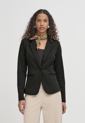 Femme aux cheveux bouclés portant un blazer noir, une écharpe beige nouée au cou et un pantalon beige clair sur un fond clair uni.