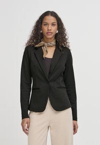 Černý přizpůsobený blazer s jedním knoflíkem, předními kapsami a slim fit střihem. Nosič má na sobě tmavý top, doplněný o světle béžové kalhoty.