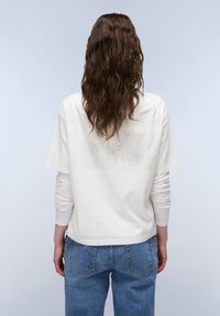 Witte T-shirt met korte mouwen en een gestructureerd ontwerp op de rug, gecombineerd met lichtblauwe hoge spijkerbroeken en lange, golvende bruine haren.