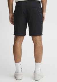 Svarte shorts laget av teksturert stoff, med en brettet hem, to baklommer med knapper, og brukt med hvite sokker og sneakers.