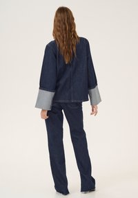 Asuste denim terdiri dari kemeja oversized biru tua dengan aksen biru muda di ujung lengan dan celana model lurus yang sesuai dengan ujung yang berbulu.