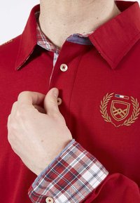 T-shirt polo rouge avec un col à carreaux bleu et blanc. Comprend un logo brodé et des boutons sur la patte de boutonnage et les poignets, en tissu coton.