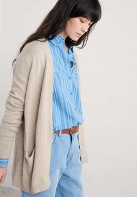 Bežs adīts kardigāns ar garām piedurknēm un kabatām, pār kādu zilu svītrainu krekliņu ar pogām. Gaiši zili augstiem jostām izgriezti džinsi ar brūnu jostu.