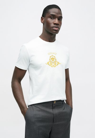 Hvid bomuld T-shirt med et gyldent Versace-logo på forsiden. Modellen bærer koksgrå bukser med hænderne i lommerne.