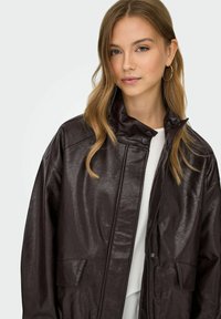 Giacca in pelle marrone con colletto alto, chiusura a pressione e due tasche frontali; presenta una texture lucida e una vestibilità comoda.