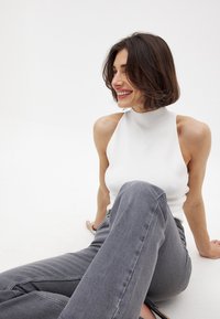 Valkoinen hihaton paita, jossa on korkea kaula-aukko, yhdistettynä korkeavyötäröisiin harmaisiin farkkuihin. Kangas näyttää sileältä ja istuvalta. Yksinkertainen muotoilu, ilman kuvioita.