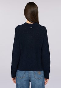 Granatowy sweter z dzianiny o teksturowanym wzorze w jodełkę, z prążkowanym dekoltem i małą metką marki z tyłu. Pokazany z niebieskimi dżinsami.