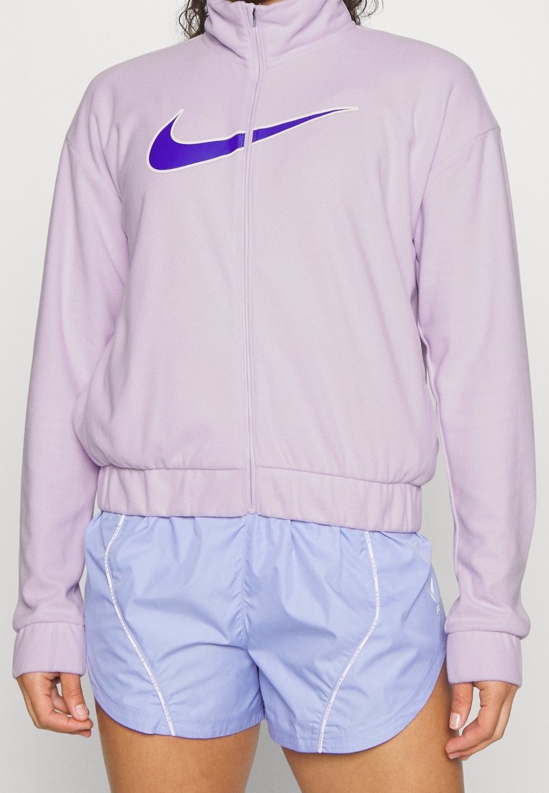 Lila vetoketjupaita, jossa on valkoinen ja violetti Nike-logo. Yhdistetty vaaleansinisiin urheilushortseihin, joissa on valkoinen reuna ja joustava vyötärö.