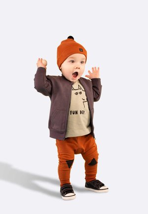 Braune Übergangsjacke mit Reißverschluss, olivfarbenes T-Shirt mit Katzenmotiv, orangefarbene Jogginghose mit schwarzen Flecken und schwarze Sneakers mit weißen Sohlen.