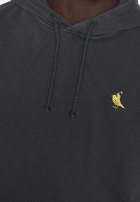 Sudadera con capucha azul marino hecha de algodón, con capucha ajustable mediante cordón y un pequeño bordado de un pájaro dorado en el pecho izquierdo. Textura suave.