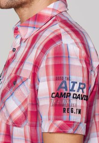 Rot- und blau kariertes Kurzarmhemd mit Button-Down-Kragen, das einen Grafikdruck auf dem Ärmel und eine Brusttasche aufweist.