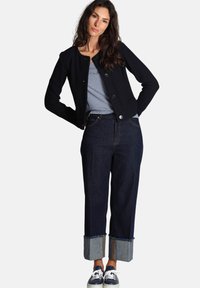 Giacca cropped blu scuro con bottoni sopra una maglietta a righe, abbinata a jeans a gamba larga in denim scuro con orli risvoltati e sneakers blu.