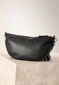 Sac bandoulière en cuir noir avec fermoirs argentés et une bandoulière réglable rayée noir et blanc sur un piédestal en pierre.