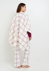 Poncho a quadri bianco con linee bordeaux, realizzato in tessuto morbido; indossato sopra pantaloni abbinati a quadri bianchi, presenta un design ampio e fluente.