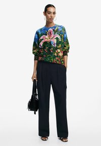 Maglione a fantasia floreale con colori vivaci, colletto blu; abbinato a pantaloni scuri e comodi e borsa a tracolla nera.