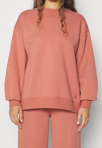Donna che indossa una felpa oversized rosa polvere e pantaloni abbinati, con i capelli sciolti e le mani rilassate ai lati.