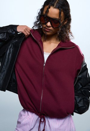 Persoană cu păr creț purtând ochelari de soare mari și întunecați, o jachetă bordo cu fermoar și mâneci din piele neagră și pantaloni roz cu dungi.