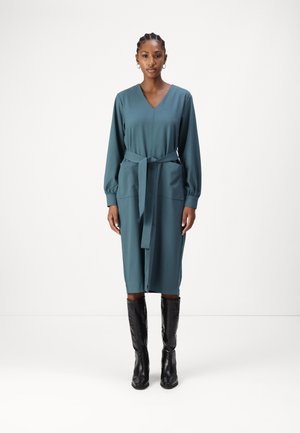 Robe teal, longueur genou, avec de longues manches bouffantes et un décolleté en V. Elle possède une ceinture en tissu et deux poches avant. Portée avec des bottes noires jusqu'au genou.