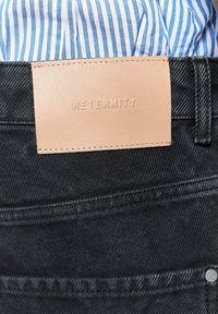 Toppa in pelle con la scritta "RETERNITY" impressa su jeans di denim neri indossati con una camicia a righe blu e bianca infilata dentro.