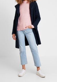 Cappotto blu navy a trama, maglione rosa, jeans cropped azzurri chiari e sneaker bianche. Il cappotto presenta un cappuccio e dettagli con bottoni.