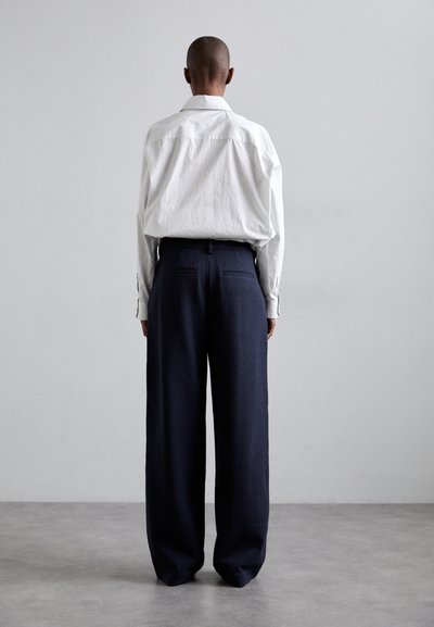 Camicia bianca a maniche lunghe con vestibilità rilassata, infilata in pantaloni larghi blu scuro con una sottile texture e tasche posteriori.