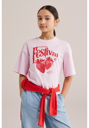 Persoană tânără purtând un tricou roz „Strawberry Festival” cu un grafic cu căpșuni roșii, pulover roșu legat în jurul taliei și pantaloni albastru deschis.