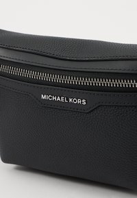 Černá koženková taška s texturou, stříbrným zipem a štítkem s nápisem "MICHAEL KORS." Vyznačuje se hladkým, zaobleným tvarem.