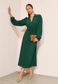 Dlhé zelené šaty s golierovým výstrihom, sčisteným pásom a voľnými rukávmi, v kombinácii s hnedou kabelkou a topánkami so vzorom hada.