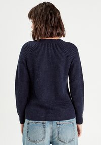 Maglione a maglia di un blu navy scuro con texture a coste e maniche lunghe, dotato di un collo rotondo e un orlo aderente, abbinato a jeans azzurri chiari.