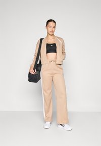 Casaco desportivo bege com riscas brancas, calças a condizer e um top crop preto. Inclui uma mala de ginásio preta e ténis brancos. Textura suave.