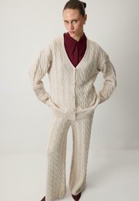 Beige kabelgebreide cardigan en bijpassende broek met een V-hals, oversized pasvorm en knoopsluiting. Donkerrode kraagshirt eronder, met een gestructureerde stof.