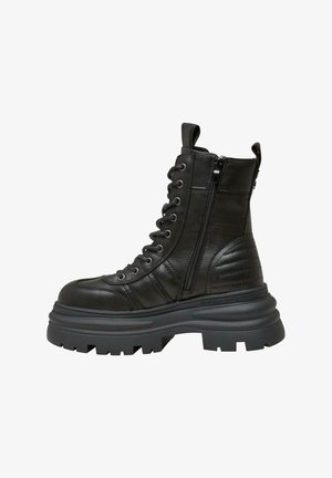 Botín de cuero negro hasta el tobillo con suela gruesa, cordones en la parte delantera y cremallera lateral. Presenta detalles acolchados en los costados.